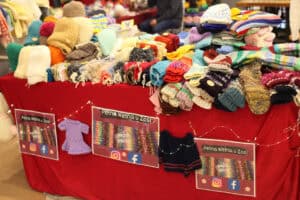 Stoisko „Pełna Wełna u Zosi”. Kolekcja kolorowych, dzianinowych rękawiczek i czapek ułożonych gęsto na czerwonym podłożu. Rękawiczki ozdobione są różnorodnymi motywami, takimi jak paski, wzory, melanż kolorów. Wyraźnie widać dbałość o detale i duży wybór wariantów kolorystycznych włóczki, od beżów po błękity, zielenie, czerwienie.