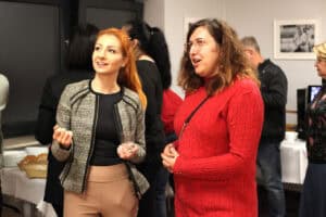 Dwie kobiety rozmawiają podczas wernisażu. Kobieta po lewej, o rudych włosach spiętych w kucyk trzyma w dłoni szklankę z wodą i z uśmiechem patrzy poza kadr. Kobieta po prawej, o kręconych włosach i w okularach, ubrana w jaskrawoczerwony sweter, również żywo rozmawia. W tle widoczne są inne osoby i stół z poczęstunkiem