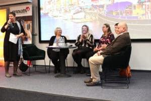 Pięcioro panelistów (dr Marta Komorska, Małgorzata Zmysłowska, Karolina Kowalczyk, Justyna Spryszak, Dorota Krać, Wojciech Dec) uczestniczy w konferencji na scenie. Po lewej stronie stoi kobieta w czarnej sukience i niebieskim wzorzystym szalu (dr Marta Komorska), która przemawia do mikrofonu, trzyma w dłoni notatki. Pozostałe cztery osoby (trzy kobiety i jeden mężczyzna) siedzą na ciemnozielonych fotelach wokół małego, okrągłego stolika, na którym stoją butelki z wodą i szklanki. W tle znajduje się bardzo duży ekran wyświetlający kolorowe zdjęcie portu z cumującymi łodziami, a po lewej stronie widoczny jest baner z logo „innovativ”.