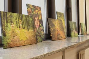 Długie ujęcie (w perspektywie zbieżnej) rzędu wydruków na płótnie, przedstawiających postacie w scenerii leśnej, ustawionych na szerokim, nakrapianym parapecie. Na pierwszym planie wydruk, na którym kobiety stoją w lesie w długich sukniach wykonanych z mchu lub liści. Obok nich znajduje się portret osoby z głową i ciałem ukrytym w liściach dębu. W tle widoczne są kolejne obrazy w tematyce leśnej, rozciągające się wzdłuż okna