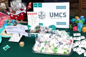 Centralne ujęcie stoiska. W tle stoi duża, biała tablica z logo UMCS i napisem „Wydział Historii i Archeologii". Przed tablicą leżą woreczki foliowe wypełnione słodyczami i małymi, zabawnymi etykietkami. Na środku widoczna jest odręczna notatka "HISTAR’owe pralinki!" z listą słodyczy. Po prawej stronie znajdują się liczne, kolorowe sześciany z logo UMCS.