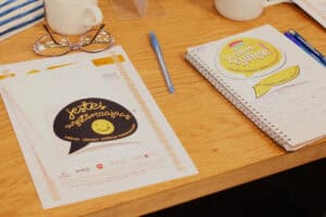 Blat stołu z materiałami warsztatowymi. Po lewej biały arkusz z grafiką dymka i napisem „Jesteś wystarczający”, powyżej różowe okulary. Po prawej żółty dymek z napisem „Jestem SUPER”