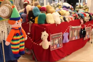 Zdjęcie przedstawia stoisko z rękodziełem. Na pierwszym planie wisi kolorowa, dziergana lalka klauna w tęczowym ubranku. W tle widać stół nakryty czerwonym materiałem, na którym piętrzą się wełniane czapki i akcesoria. Stoisko zdobią drobne światełka oraz tabliczki z nazwami mediów społecznościowych twórczyni.