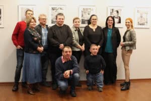 Zdjęcie grupowe dziesięciu osób na tle wystawy. Grupa stoi i kuca na jasnej, drewnianej podłodze, uśmiechając się do aparatu. Kobiety i mężczyźni ubrani są w stroje nieformalne. W tle wisi rząd czarno-białych portretów