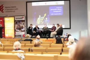 Na scenie siedzi sześcioro panelistów uczestniczących w dyskusji (dr Marta Komorska, Małgorzata Zmysłowska, Karolina Kowalczyk, Justyna Spryszak, Dorota Krać, Wojciech Dec). Po lewej stronie widoczne są wysokie banery reklamowe. W tle na ekranie widnieje napis: „Konferencja Senioralni Liderzy Dostępności, Lublin, 16 grudnia 2025 r.”. Widok z widowni.