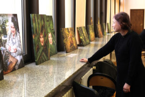 Kobieta w czarnej, dzianinowej sukience, stoi z prawej strony kadru i opiera dłoń na szerokim, nakrapianym parapecie, ogląda wystawę. Na parapecie ustawione są w rzędzie obrazy na płótnie, w tym od lewej: portret kobiety przy starej maszynie do szycia oraz dwa portrety kobiet otoczonych zielonymi liśćmi, utrzymane w kolorystyce natury. W tle widoczne są duże okna i drewniane drzwi