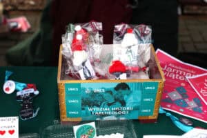 Stoisko Wydziału Historii i Archeologii. Centralny element kadru stanowi prostokątne, brązowo-turkusowe pudełko z nadrukiem z logo UMCS, nazwą Wydziału Historii i Archeologii i zdjęciem pary studentów. W pudełku znajdują się małe, ręcznie robione figurki w czapkach Mikołaja – białe i czerwone – oraz drobne słodycze i ozdoby zapakowane w celofan.