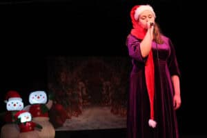 Występ muzyczny Julii Majsterek. Młoda kobieta w długiej fioletowej welurowej sukni i dużej czapce Mikołaja śpiewa do mikrofonu na ciemnej scenie, oświetlonej ciepłym światłem. Po lewej stoją dmuchane bałwany w czapkach Mikołaja i podświetlony baner z zimową, świąteczną grafiką.