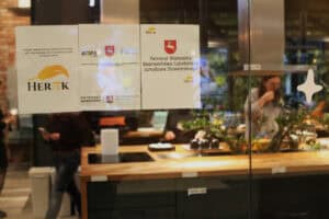 Trzy białe arkusze papieru naklejone na szybę informujące o projekcie „Matka dziecka niepełnosprawnego – siła i moc każdego dnia”, partnerach i patronatach. Przez szybę widać rozmyte wnętrze lokalu z nowoczesną kuchnią.