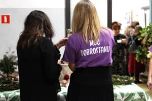 Widok od tyłu na kobietę w fioletowej koszulce „MOC DOBROSTANU” rozmawiającą z uczestniczką ubraną na czarno. Stoją przy stole przykrytym białym obrusem w zielone liście, na którym leżą wieńce adwentowe. Po prawej, w głębi widać grupę osób, które znajdują się w innej sali.