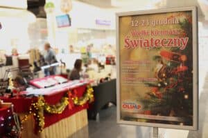 Zdjęcie przedstawia plakat informacyjny umieszczony w srebrnej ramie na stojaku, znajdujący się w pasażu handlowym. Plakat głosi: „12-23 grudnia Wielki Kiermasz Świąteczny” i zawiera logotyp Galerii Olimp. Na plakacie wymieniono dostępne produkty, takie jak żywność tradycyjna (wędliny, sery, miody, ciasta) oraz rękodzieło artystyczne (biżuteria, ozdoby świąteczne). W rozmytym tle widać fragmenty stoisk kiermaszowych udekorowanych girlandami i światełkami.