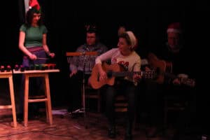 Czworo dorosłych muzyków występuje na scenie na tle ciemnej kurtyny. Po lewej Dorota Skałecka w zielonej koszulce i czapce elfa gra na kolorowych dzwonkach. W centrum Karolina Czajka w jasnym swetrze i czapce Mikołaja gra na gitarze akustycznej. Za nimi siedzą dwaj mężczyźni; jeden w okularach z opaską z porożami gra na dzwonkach, drugi w czapce Mikołaja gra na gitarze.