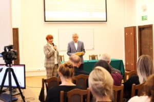 Kobieta o rudych włosach (Maria Pietrusza-Budzyńska), ubrana w jasny, kraciasty garnitur, stoi na przedzie sali, trzymając mikrofon i przemawiając do zgromadzonej publiczności. Obok niej stoi mężczyzna (prof. Wojciech Otrębski) w szarym garniturze, z żółtą teczką w ręku. Za nimi widoczny jest duży ekran projekcyjny z tekstem oraz biała tablica. W tle widoczne są również drewniane drzwi i szafka. Na pierwszym planie, z lewej strony, stoi kamera wideo na statywie, a na prawo od niej, widać tyły głów słuchaczy siedzących na drewnianych krzesłach