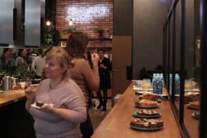 Długi jasnobrązowy stół z talerzami ciasta, butelkami wody i szklankami ustawionymi w rzędzie. Na pierwszym planie kobieta w szarej bluzce trzyma talerzyk. W tle czerwona ceglana ściana z napisem 'SZWEDZKI STÓŁ' i grupa osób rozmawiających przy meblach kuchennych.