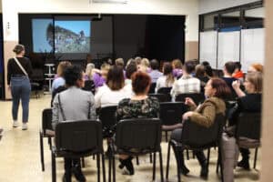 Widok na salę widowiskową podczas prezentacji projektu. Uczestnicy siedzą tyłem do obiektywu na czarnych krzesłach, patrząc w stronę ekranu projekcyjnego. Na ekranie widoczne jest zdjęcie grupy uśmiechniętych kobiet w plenerze. Po lewej stronie sali stoi kobieta w ciemnym ubraniu, a w głębi widać przygotowane do występu instrumenty etniczne oraz gongi.