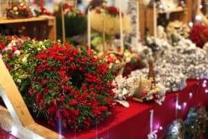 Stoisko handlowe z rękodziełem bożonarodzeniowym. Na pierwszym planie od lewej widać gęsty, okrągły stroik wykonany z zielonego igliwia i jaskrawoczerwonych suszonych kwiatów. W tle, na czerwonym obrusie, wyeksponowane są liczne dekoracje: srebrzyste wianki, białe świece oraz stroiki ozdobione bombkami. Całość oświetlona jest ciepłym światłem lampek choinkowych.