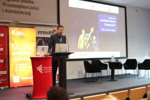 Zbliżenie na prelegenta (Jacek Lis) przemawiającego zza mównicy na scenie. Mężczyzna w ciemnej marynarce i koszuli mówi do mikrofonu, patrzy w stronę publiczności. Obok niego, na czerwonej osłonie mównicy, widoczne jest logo uczelni WSPA. W tle znajduje się duży ekran z grafiką konferencyjną przedstawiającą seniorów oraz tytuł wydarzenia: „Senioralni Liderzy Dostępności”. Po prawej stronie sceny stoją puste, eleganckie, czarne fotele i stolik z wodą.