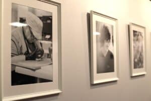 Trzy czarno-białe fotografie w białych, prostokątnych ramach, wiszące w rzędzie na jasnej ścianie. Na pierwszym planie (po lewej) widać wyraźnie ujęcie osoby w czapce, pochylonej nad blatem i intensywnie pracującej rękami. Dwa pozostałe zdjęcia (po prawej) są rozmyte; środkowe przedstawia uśmiechniętą osobę z bujnymi, kręconymi włosami