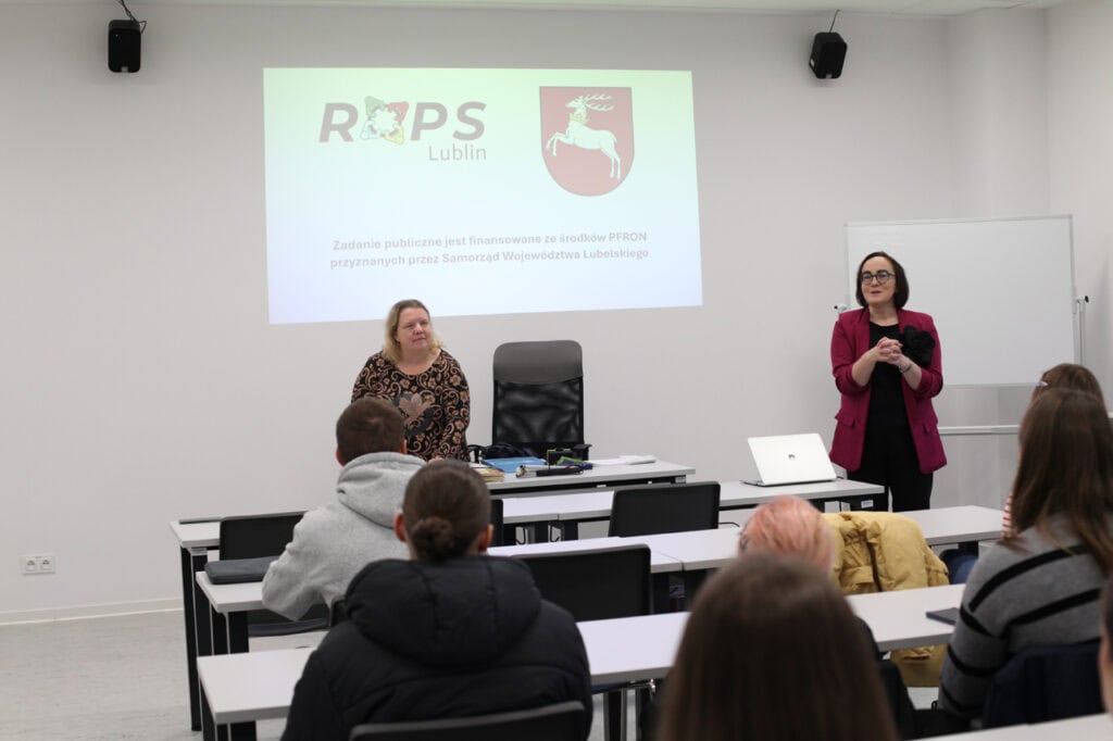 Wnętrze sali wykładowej. Od lewej kobieta (Ewelina Orzechowska) w ciemnej, wzorzystej bluzce stoi za biurkiem i przemawia do grupy młodych ludzi siedzących w rzędach białych ławek. Z prawej kobieta (Anna Bieganowska-Skóra) w marynarce w kolorze fuksji i czarnych spodniach stoi i gestykuluje w stronę osób siedzących w ławkach. Na pierwszym planie widać uczestników szkolenia, siedzących tyłem do obiektywu. Na tle przeciwległej, jasnej ściany widnieje logo "ROPS Lublin" oraz informacja o finansowaniu z PFRON