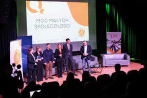 Sześć osób (trzech mężczyzn i trzy kobiety) stoi na scenie obok siebie. Wszyscy ubrani są w stroje biznesowe. W tle widoczny jest ekran z hasłem "Moc Małych Społeczności" i trzy szare kanapy. Na pierwszym planie, w dolnej części kadru, siedzi w rzędach publiczność