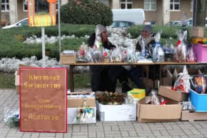 W lewym dolnym rogu duża, pionowa tablica w kolorze brązowym, oprawiona czerwoną ramką, z wyraźnym napisem „Kiermasz Świąteczny Dom Środowiskowy Roztocze", stoi na pierwszym planie na kostce brukowej. Za drewnianym stołem siedzą dwie uśmiechnięte kobiety w ciepłych, zimowych czapkach. Stół jest zastawiony licznymi, zapakowanymi paczkami świątecznymi przewiązanymi kokardami, a pod stołem stoją kartonowe pudła z paczkami i prezentami.