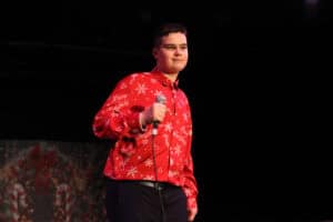 Występ sceniczny Tymoteusza Misiury. Młody, uśmiechnięty artysta, ubrany w czerwoną koszulę z białymi świątecznymi wzorami (płatki śniegu i napisy „Merry Christmas") oraz ciemne spodnie. Trzyma mikrofon w prawej ręce. Jego wzrok skierowany jest lekko w prawą stronę kadru. Tło jest czarne, a w lewym dolnym rogu widoczny jest niewyraźny fragment dekoracji świątecznej w kolorach zieleni i czerwieni.