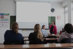 W jasnej sali wykładowej, Anna Bieganowska-Skóra i Dorota Krać znajdują się przy biurku i są pogrążone w rozmowie. Na pierwszym planie widać słuchaczy wykładu, siedzą tyłem do obiektywu. Jedna z prowadzących siedzi, obsługuje laptopa, druga stoi. W tle na ścianie duży, biały ekran rzutnika