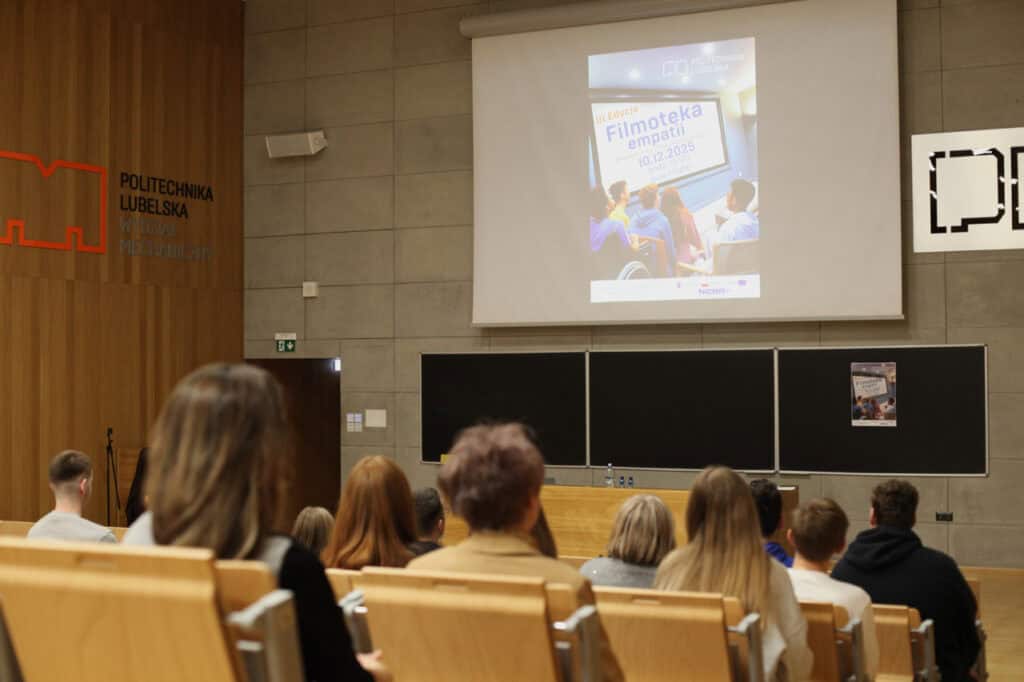 Sala wykładowa Wydziału Mechanicznego Politechniki Lubelskiej podczas wydarzenia. Uczestnicy siedzą w drewnianych ławach, zwróceni tyłem do obiektywu. W centralnym punkcie znajduje się duży ekran projekcyjny, na którym wyświetlana jest grafika promująca III edycję „Filmoteki empatii”. Na lewej ścianie, pokrytej drewnianymi panelami, umieszczono pomarańczowy logotyp uczelni i nazwę wydziału
