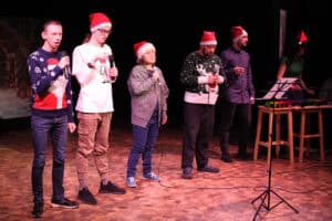 Czterech młodych mężczyzn i jedna kobieta stoją na scenie w czapkach Mikołaja i śpiewają do mikrofonów. Po lewej mężczyźni w granatowym swetrze i białej koszulce. W centrum kobieta w dużej szarej bluzie. Po prawej dwaj mężczyźni w zielonym swetrze i ciemnej koszuli. W prawym rogu przy statywie z dzwonkami stoi kobieta w zielonej koszulce.