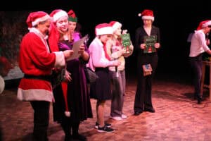 Grupa osób stoi na drewnianej scenie w strojach świątecznych i trzyma dyplomy. Po lewej mężczyzna w stroju Mikołaja, obok kobieta w fioletowej sukni i długiej czapce świątecznej. W tle widoczna postać w zielonym stroju elfa. Trzy kobiety trzymają zielone dyplomy, a po prawej w głębi kobieta w białej koszuli mówi do mikrofonu.