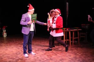 Dwaj mężczyźni stoją na drewnianej scenie podczas wręczania dyplomów. Po lewej mężczyzna w szarej marynarce i okularach trzyma zielony dyplom. Po prawej mężczyzna w stroju Mikołaja trzyma dużego skrzata w złotej czapce. Za nim stoi kobieta w świątecznej czapce. Na scenie widać stołek z kolorowymi instrumentami perkusyjnymi i w lewym dolnym rogu dmuchanego pingwinka w czapce Mikołaja.