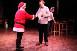 Ceremonia wręczania upominków na scenie. Mężczyzna w stroju Mikołaja ściska dłoń mężczyzny w białej koszuli, który trzyma białą kartkę lub dyplom. Po prawej za stołem stoi kobieta w jasnej koszulce i czapce Mikołaja. Po lewej widać fragment dekoracji ze świecącym pingwinem w świątecznej czapce.