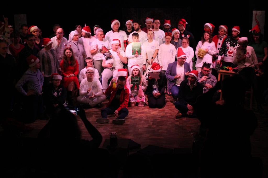 Grupa kilkudziesięciu osób stoi i siedzi na drewnianej scenie do pamiątkowego zdjęcia. Większość ma świąteczne nakrycia głowy, a część białe koszulki i czerwone elementy garderoby. Wiele osób trzyma dyplomy. Na pierwszym planie widać zarys głowy i rąk widza. Scena jest oświetlona, tło ciemne.