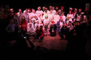 Grupa kilkudziesięciu osób stoi i siedzi na drewnianej scenie do pamiątkowego zdjęcia. Większość ma świąteczne nakrycia głowy, a część białe koszulki i czerwone elementy garderoby. Wiele osób trzyma dyplomy. Na pierwszym planie widać zarys głowy i rąk widza. Scena jest oświetlona, tło ciemne.