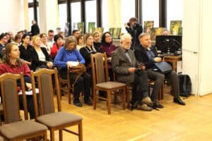 Zgromadzeni uczestnicy konferencji siedzą na drewnianych krzesłach w sali z jasną, drewnianą podłogą i dużymi oknami w tle. W środku kadru widoczny jest starszy mężczyzna z brodą (prof. Piotr Oleś), w tweedowej marynarce, trzymający ulotkę. Z tyłu widać kilka dużych wydruków/obrazów opartych o ramy okienne