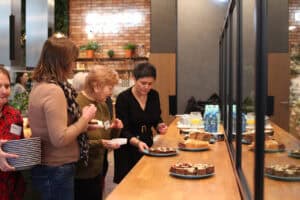 Grupa kobiet przy drewnianym blacie. Kobieta w czerwonej koszuli trzyma stos talerzy, obok inne rozmawiają. Ostatnia kobieta kładzie błękitny talerz z ciastem. Po prawej pionowe czarne ramy przeszkleń, w tle ceglana ściana z zielonymi roślinami i dekoracyjnymi żarówkami