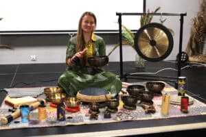 Uśmiechnięta kobieta w zielonej sukience siedzi na kolorowej macie na podeście, otoczona instrumentami relaksacyjnymi. Trzyma w dłoniach misę tybetańską i pałeczkę. Przed nią i obok niej rozłożone są liczne misy dźwiękowe o różnych rozmiarach, dzwonki koshi, bęben językowy (tank drum) oraz inne instrumenty perkusyjne. W tle po prawej stronie widać duży gong zawieszony na statywie oraz dekoracje z suszonych traw.