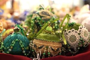 Ręcznie robione bombki choinkowe o bogatych zdobieniach. Bombki wykonane są z satynowych wstążek w odcieniach zieleni, oliwki i turkusu. Ozdobiono je koronkami, koralikami, cekinami oraz błyszczącymi tasiemkami. Niektóre posiadają doczepione sznury zielonych paciorków i kokardy. Ozdoby leżą na czerwonym podłożu, a tło jest rozmyte.