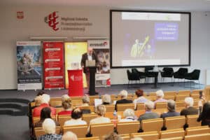 Ujęcie z tyłu sali wykładowej przedstawiające prelegenta (Wojciech Dec) – mężczyznę w brązowej marynarce, który stoi przy mównicy i korzysta z laptopa. Scena jest oświetlona, otoczona banerami informacyjnymi o studiach i projektach społecznych. Na ekranie głównym wyświetlana jest prezentacja z logo organizatorów (FLOP, WSPA). Wypełnione przez uczestników rzędy drewnianych krzeseł tworzą pierwszy plan.