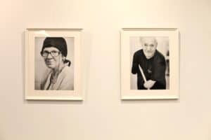 Dwa portrety w białych ramach, wiszące obok siebie na jasnej ścianie. Po lewej stronie jest czarno-białe ujęcie kobiety w okularach i czarnej chuście na głowie, patrzy prosto na widza. Po prawej stronie wisi czarno-biały portret uśmiechniętego mężczyzny w czarnej koszulce, z widoczną niepełnosprawnością rąk