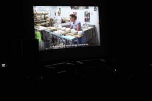 Zaciemniona sala kinowa. Na dużym ekranie wyświetlony jest kadr z filmu dokumentalnego, przedstawia dwie kobiety w pracowni rzemieślniczej i stojące w rzędach gliniane miski. Na dole ekranu widoczny jest napis w języku angielskim "Civil society also means social cooperatives" (tłum.: Społeczeństwo obywatelskie, to także spółdzielnie socjalne) oraz logo Komitetu do Spraw Pożytku Publicznego