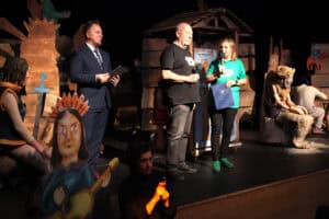 Na ciepło oświetlonej scenie teatralnej trwa przedstawienie z ludową scenografią. Na podwyższeniu stoją cztery osoby: mężczyzna w garniturze z czarną teczką, mężczyzna w czarnej koszulce i dżinsach, kpt. Tomasz Patyra, oraz Sylwia Góźdź w zielonej koszulce, trzymająca mikrofon i niebieski dokument. Po lewej siedzi postać w brązowym stroju z mieczem oraz kartonowa wycinanka postaci z gitarą i pióropuszem. Po prawej, w cieniu, widoczna jest rzeźbiona postać w wieńcu.