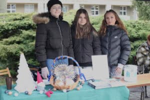 Trzy młode kobiety (studentki) w czarnych, puchowych kurtkach stoją obok siebie za stołem. Na stole prezentują ręcznie wykonane dekoracje świąteczne, w tym dużą, stożkową, białą choinkę ze sznurka, szarego krasnala w czerwonej czapce i wiklinowy kosz wypełniony ozdobami z papieru. W tle widoczne są zielone, przycięte krzewy i fragmenty budynku.
