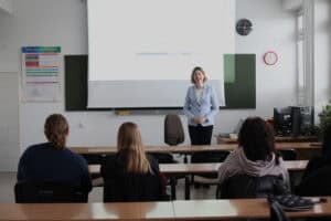 Dorota Krać prowadzi wykład w sali. Stoi pod ekranem projektora, ubrana w jasnoniebieski żakiet i czarne spodnie. Kobieta ma złożone ręce przed sobą, na wysokości bioder. Przed nią siedzą uczestnicy warsztatów, widoczni od tyłu.