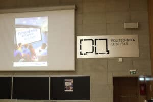 Frontowa ściana auli wykładowej Politechniki Lubelskiej. Po lewej stronie widoczny jest fragment dużego ekranu z wyświetlonym plakatem „III Edycji Filmoteki empatii”. Po prawej stronie, na ścianie z szarych płyt, znajduje się duża srebrna tablica z czarnym logotypem i napisem „POLITECHNIKA LUBELSKA”.