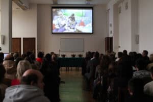 Sala wykładowa. Licznie zgromadzona publiczność siedzi w rzędach zwrócona w kierunku dużego ekranu projekcyjnego. Na ekranie wyświetlany jest kadr z filmu lub wideo, przedstawiający dwie młode kobiety siedzące obok siebie na kanapie. Poniżej ekranu znajduje się pusta, szara tablica oraz stół konferencyjny przykryty zielonym obrusem. Po obu stronach widoczne są ciemne, drewniane drzwi