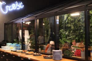 Widok przez przeszkloną czarną konstrukcję przypominającą szklarnię. W środku gęste zielone rośliny, w tym wysoki kaktus. Po lewej talerze, szklanki, butelki wody i stos talerzy. Nad ramą fragment białego neonu z napisem ‘Cześć’. Przez szyby widać rozmyte sylwetki uczestniczek siedzących przy stołach wewnątrz sali.