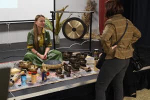 Kobieta w zielonej sukience siedzi na macie z instrumentami i rozmawia z uczestniczką wydarzenia, która stoi przed nią tyłem do obiektywu. Scena jest wypełniona misami dźwiękowymi i akcesoriami muzycznymi. W tle widać biały ekran projekcyjny oraz metalowy stojak z dużym gongiem.