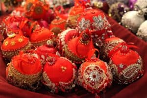 Kilkanaście ręcznie robionych bombek choinkowych w intensywnym czerwonym kolorze. Każda bombka jest bogato zdobiona złotymi haftami, cyrkoniami, białymi koralikami oraz czerwonymi wstążkami. Ozdoby leżą blisko siebie na bordowym tle, tworząc błyszczącą kompozycję.