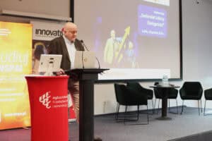 Przy mównicy stoi Wojciech Dec. Mężczyzna ma poważny wyraz twarzy i spogląda w stronę ekranu lub publiczności. Na pierwszym planie widoczna jest czerwona osłona mównicy z białym logo Wyższej Szkoły Przedsiębiorczości i Administracji. W tle widać fragment ekranu projekcyjnego oraz puste fotele na scenie.